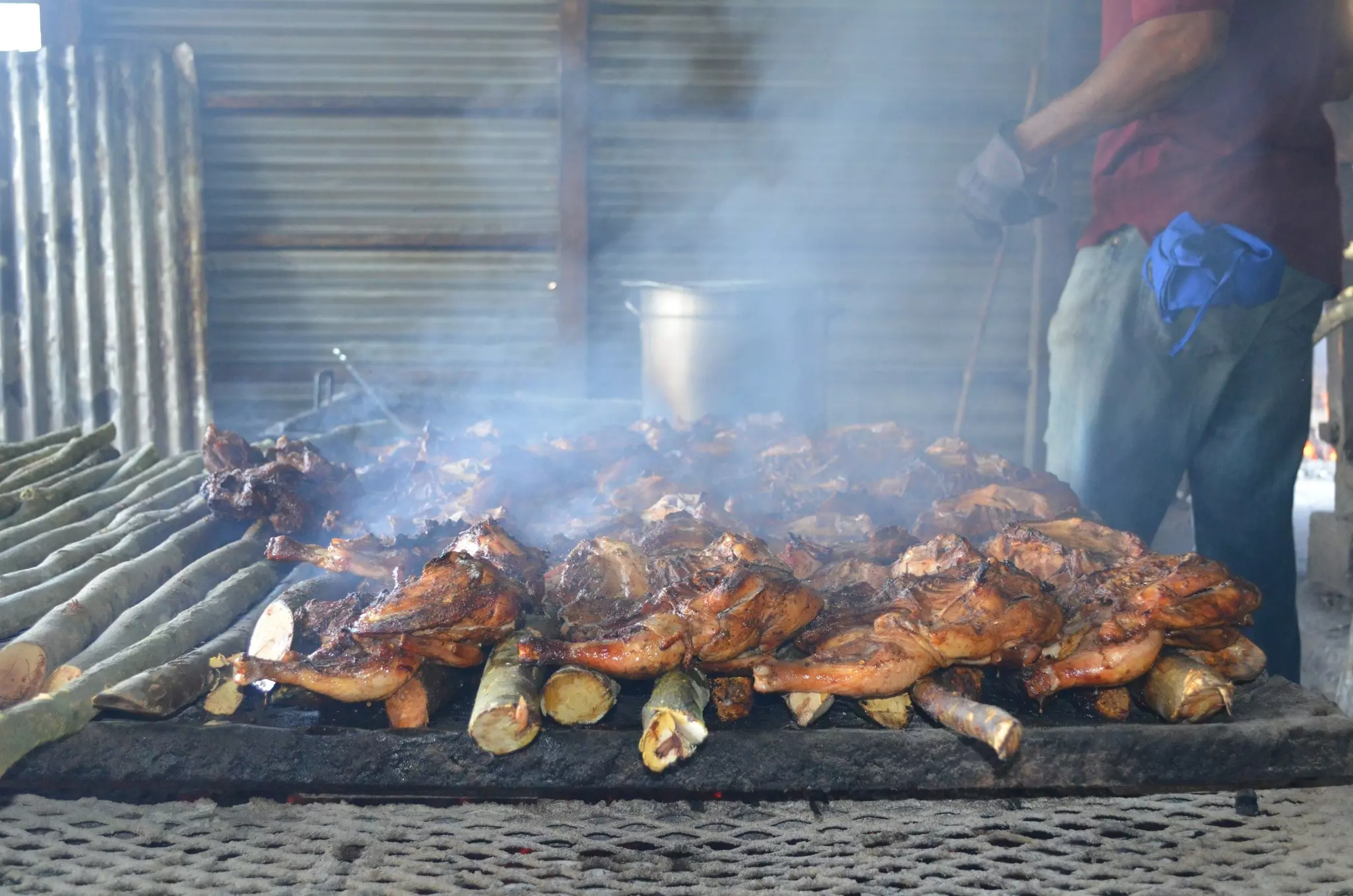 A chef stands near a charcoal fire with whole chickens on the grill and smoke rising above.