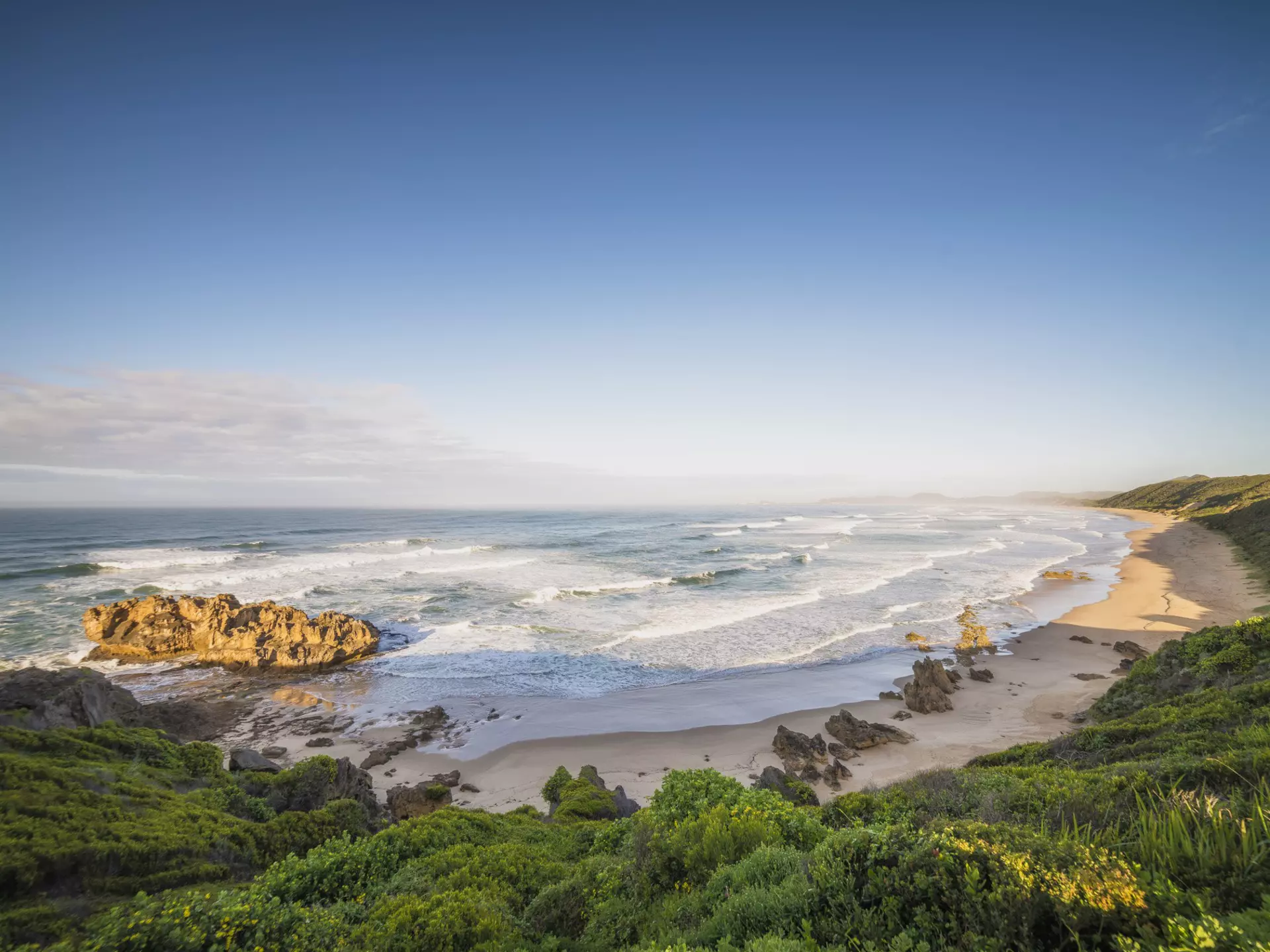 A long curve of sand backed by lush hills is pounded by waves.