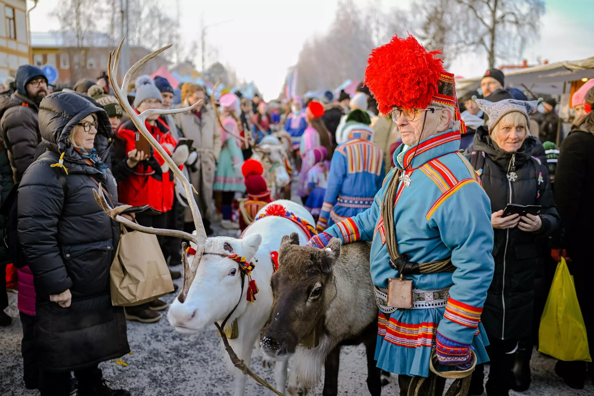 A man in traditional clothing pets a reindeer at a festival. People in heavy coats and hats surround the man and the animals.
