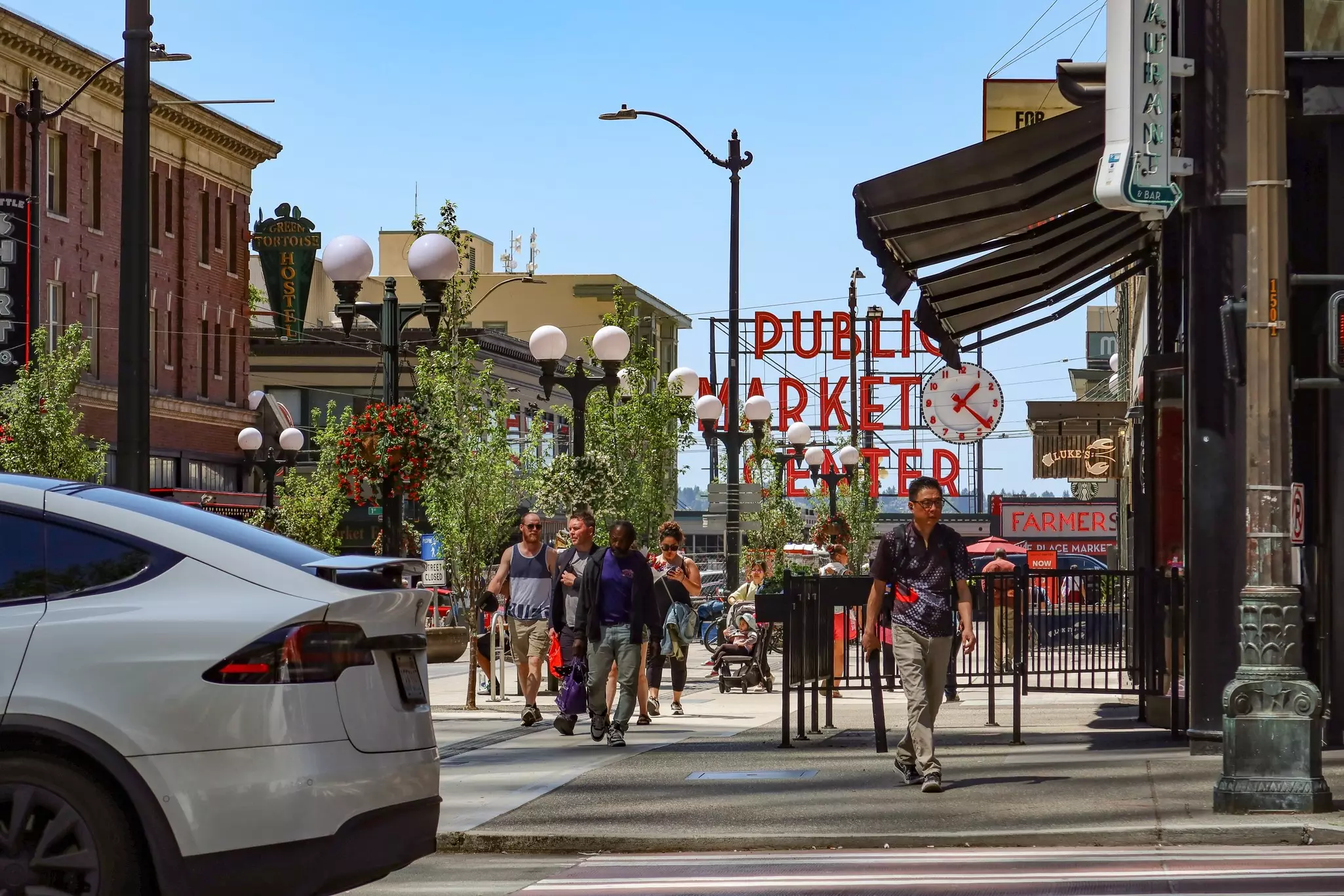 Iconic Pike Place Market sign welcomes visitors on a sunny Seattle afternoon. Pedestrians stroll past historic lamp posts and hanging flower baskets in downtown.