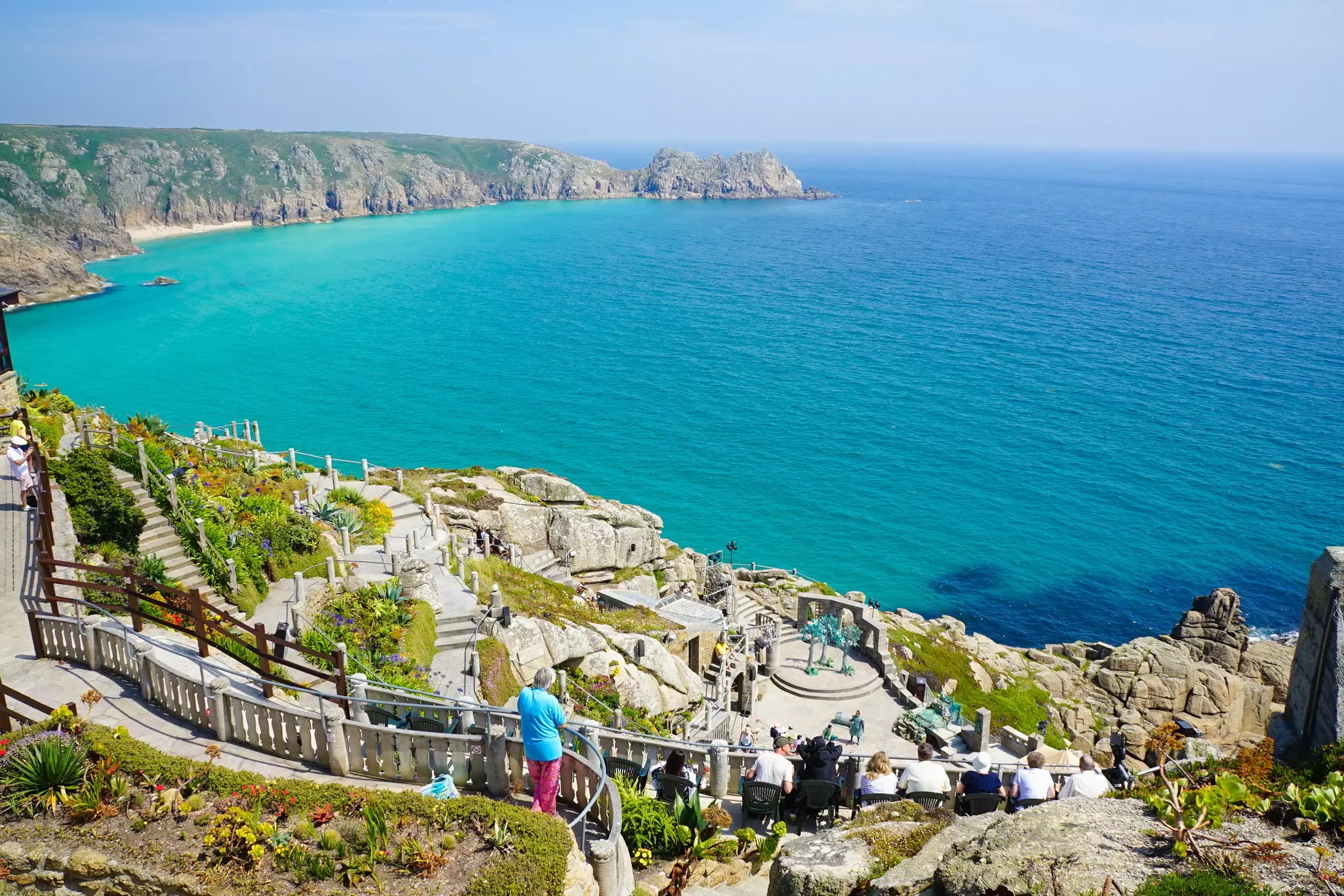 An open-air amphitheater at the edge of a cliff with vast views over the ocean and a sandy beach in the distance.  