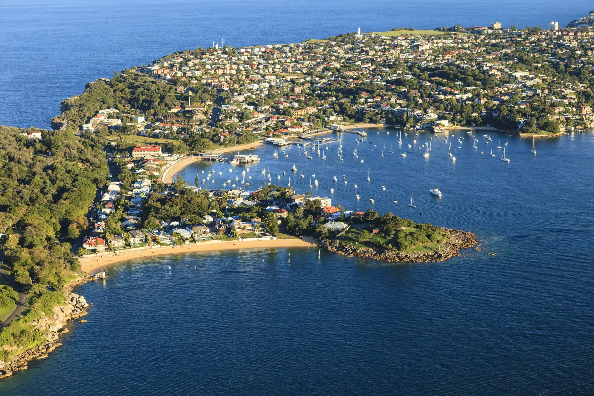 Aerial shot of bay with forested town on a spit of land and the ocean beyond on a sunny day.