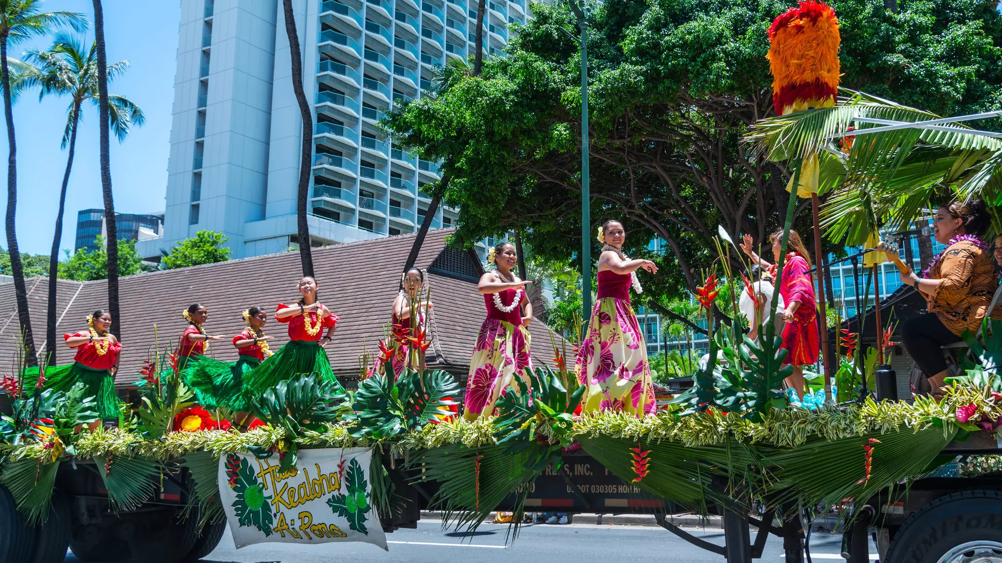 Women dancing hula on a parade float adorned with palm fronds on a sunny day.