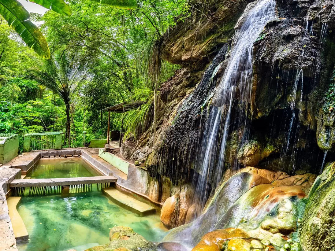 Waterfall over mineral-stained rocks into a green thermal bathing pool in rainforest, Soufrière, St Lucia