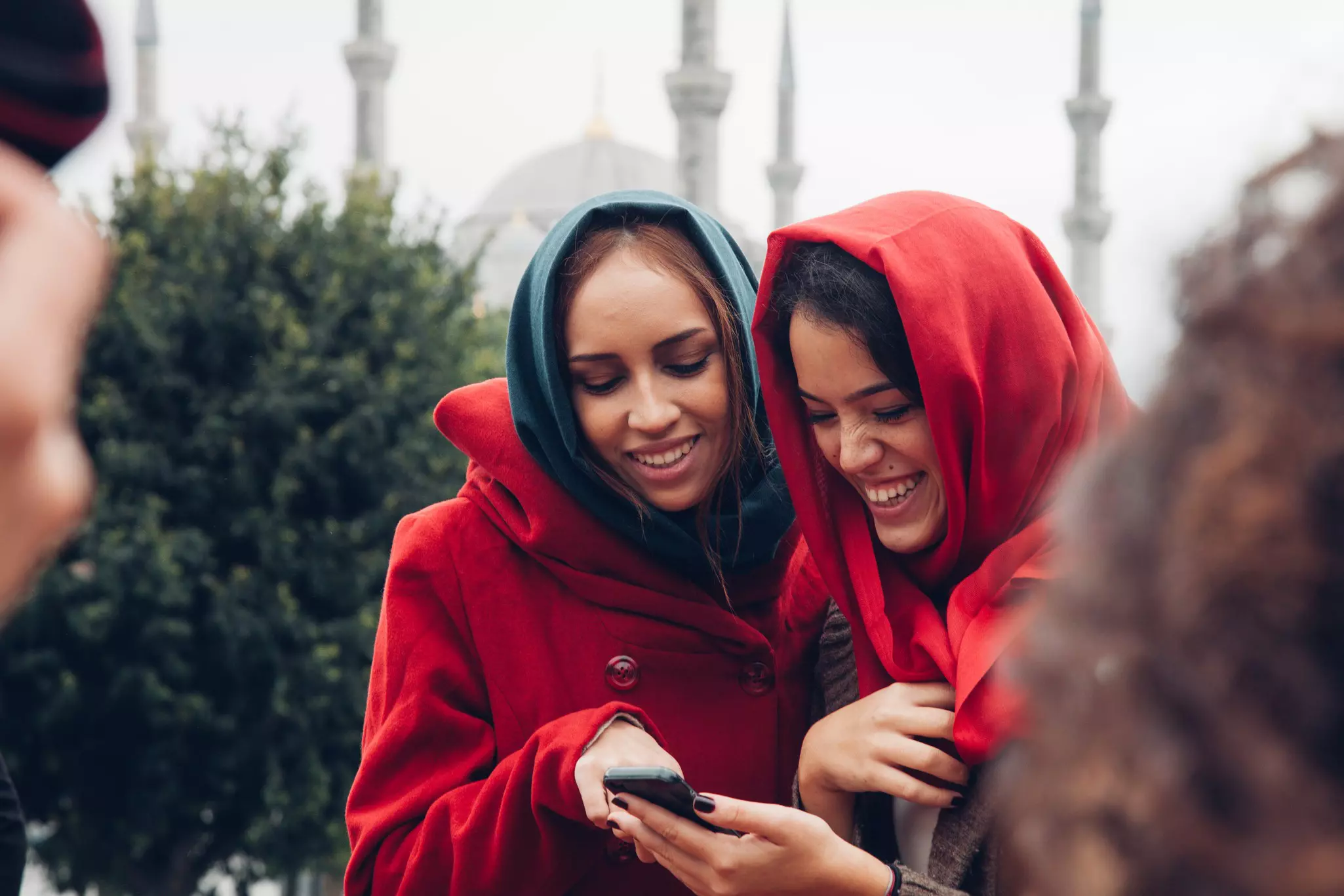 Two young women in head coverings laugh as they look at a phone near the Blue Mosque in Istanbul, Türkiye