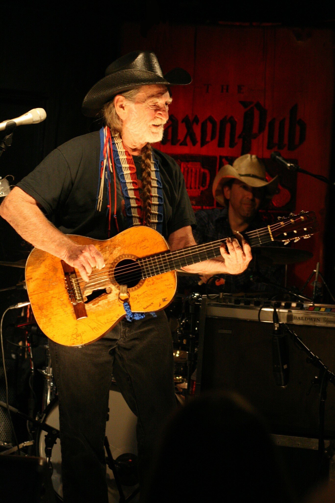 A musician wearing a black cowboy hat, black clothes and a red, white and blue accessory hanging from his neck plays a guitar.