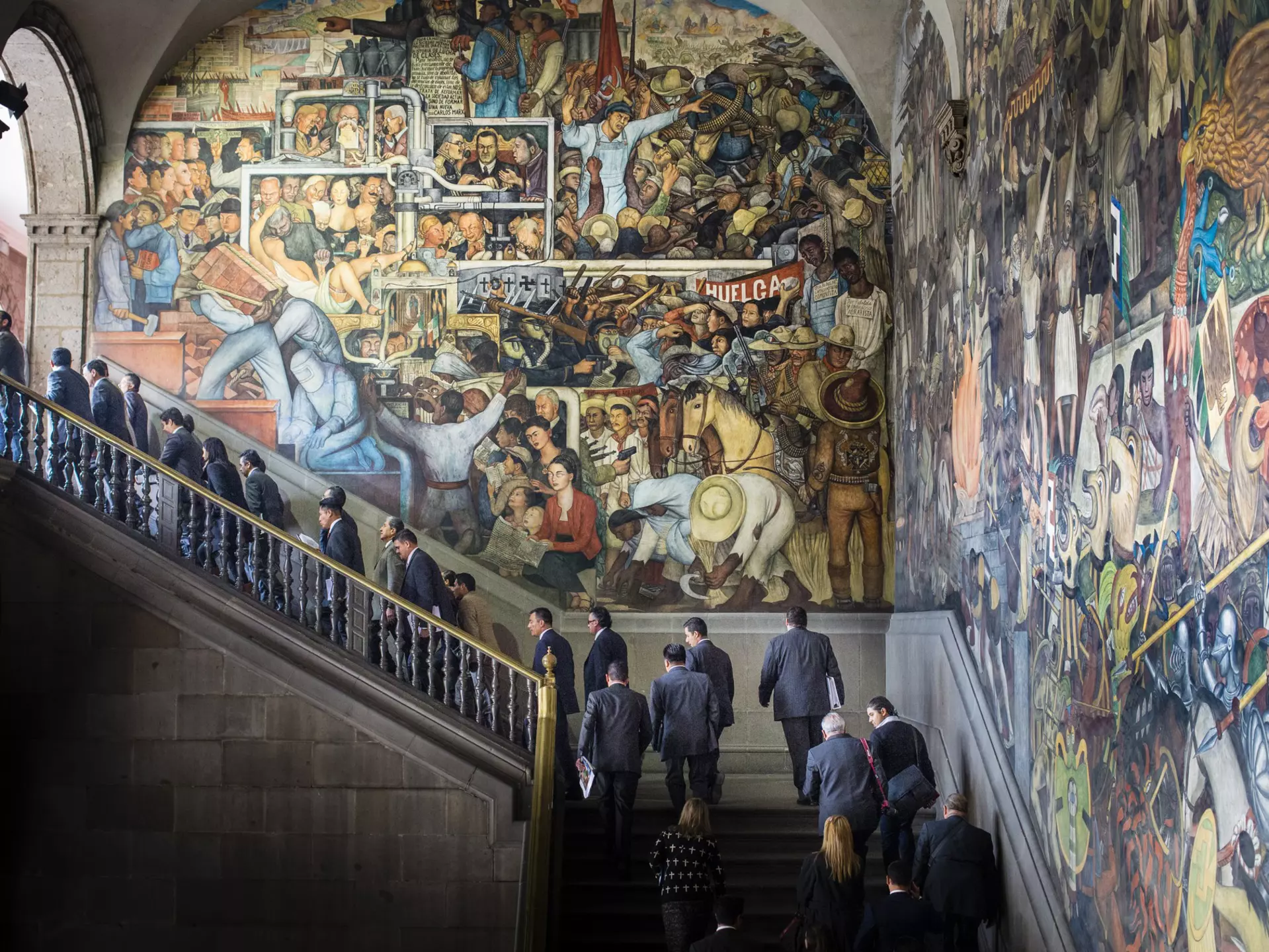 People walk up a staircase in front of a huge, elaborate, colorful mural.