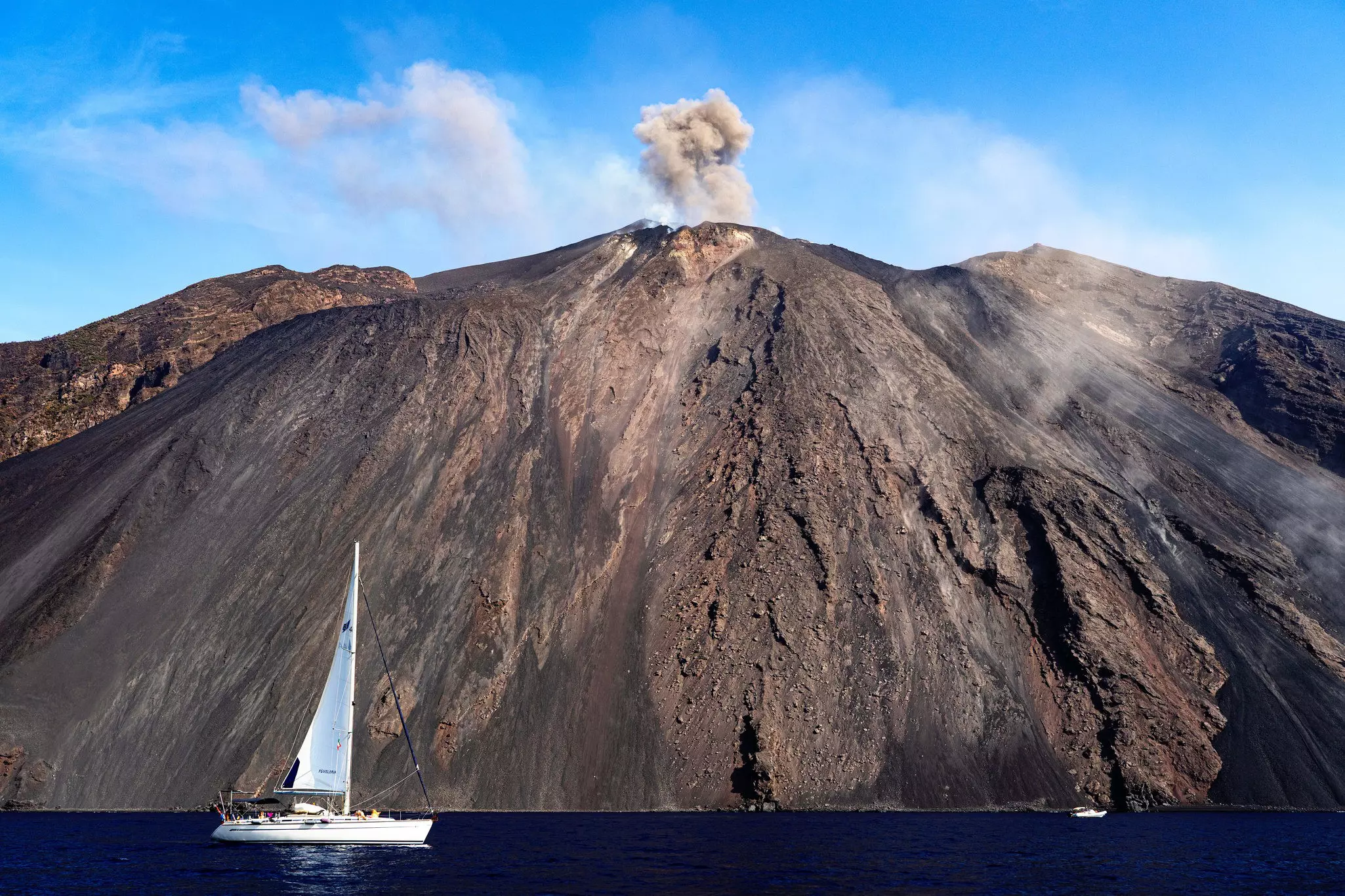 A sail boat anchored near a volcanic island where a massive wall of volcani rock and debris meets the ocean. Smoke and steam pump out the still-active volcano above.