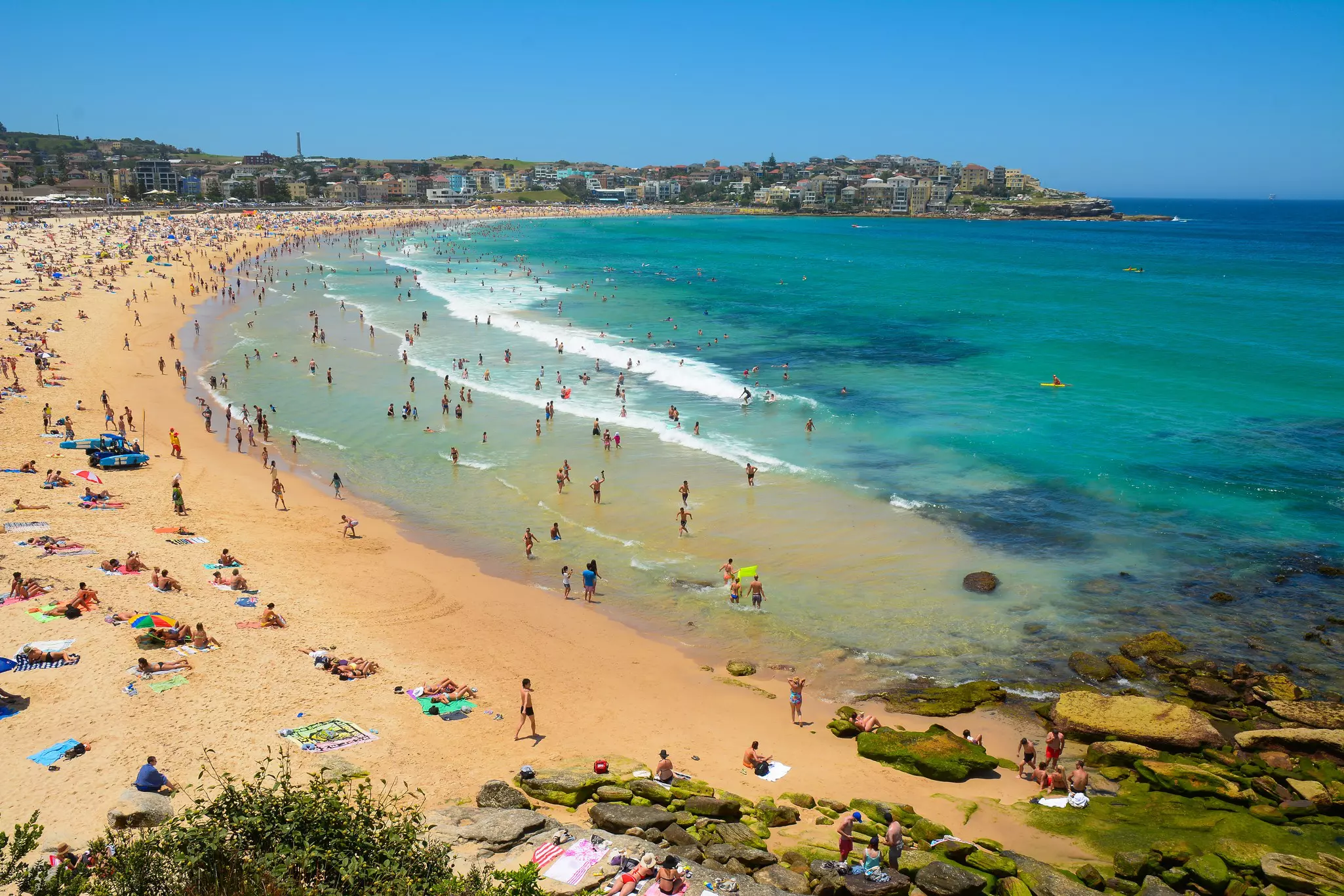 An aerial view of a crescent-shaped beach packed with people on the sand and in the water.