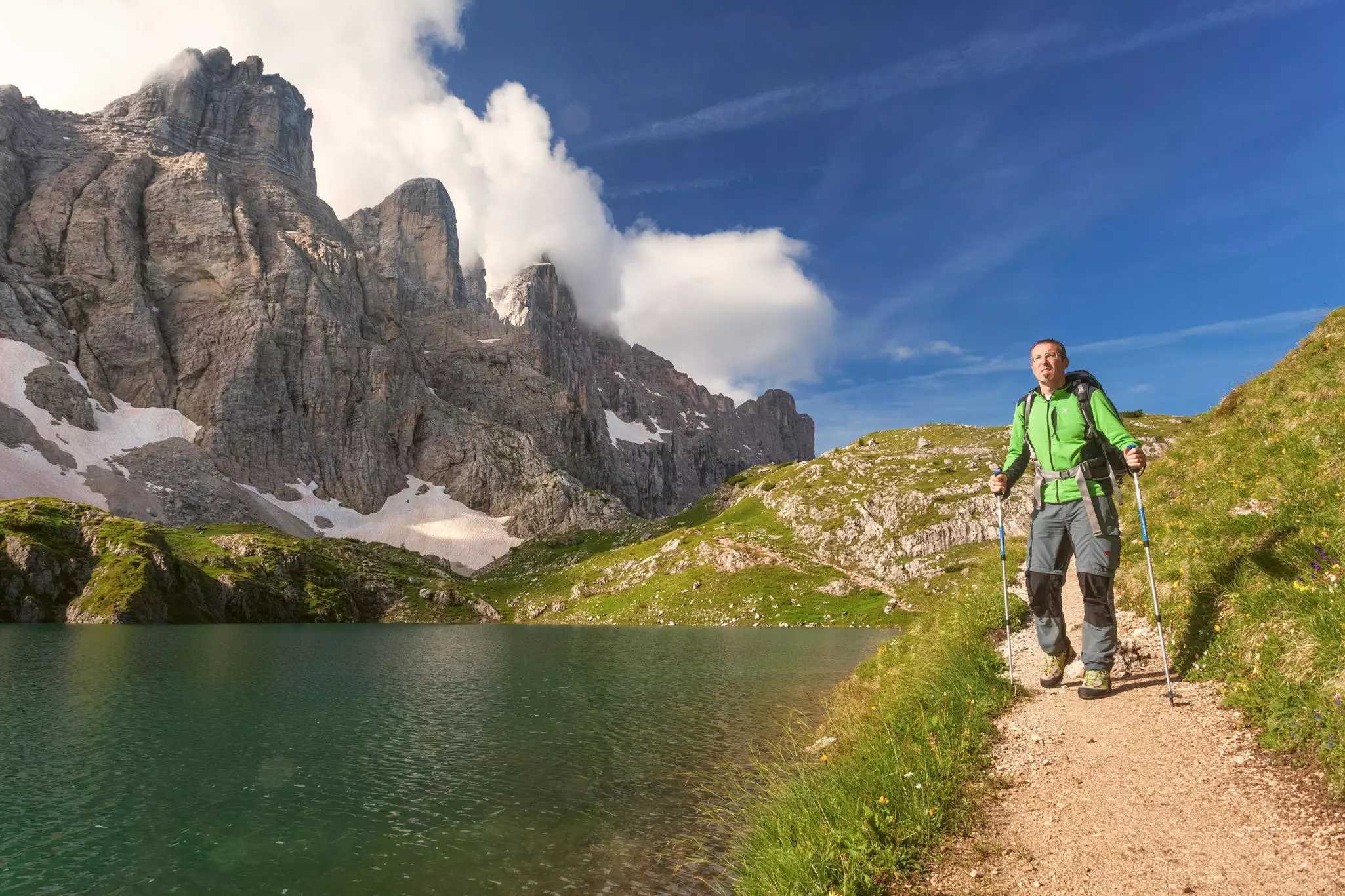 A male hiker in a green parka with poles walks along a path by a mountain lake, with steep rock peaks rising to the right