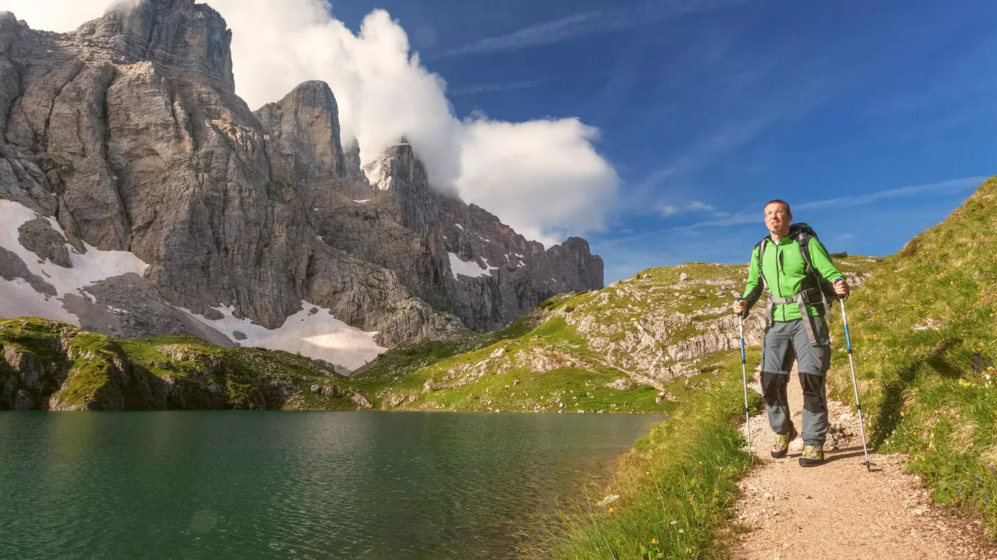 A male hiker in a green parka with poles walks along a path by a mountain lake, with steep rock peaks rising to the right