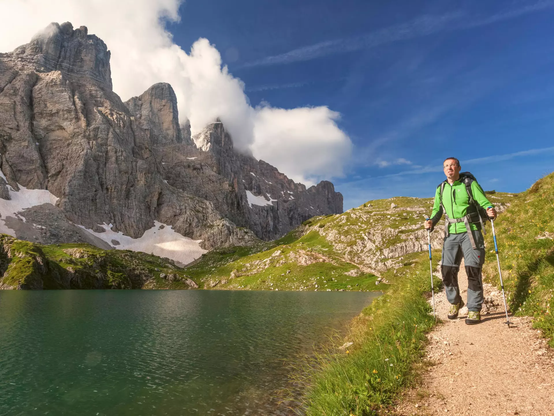 A male hiker in a green parka with poles walks along a path by a mountain lake, with steep rock peaks rising to the right