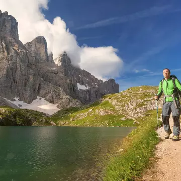 A male hiker in a green parka with poles walks along a path by a mountain lake, with steep rock peaks rising to the right