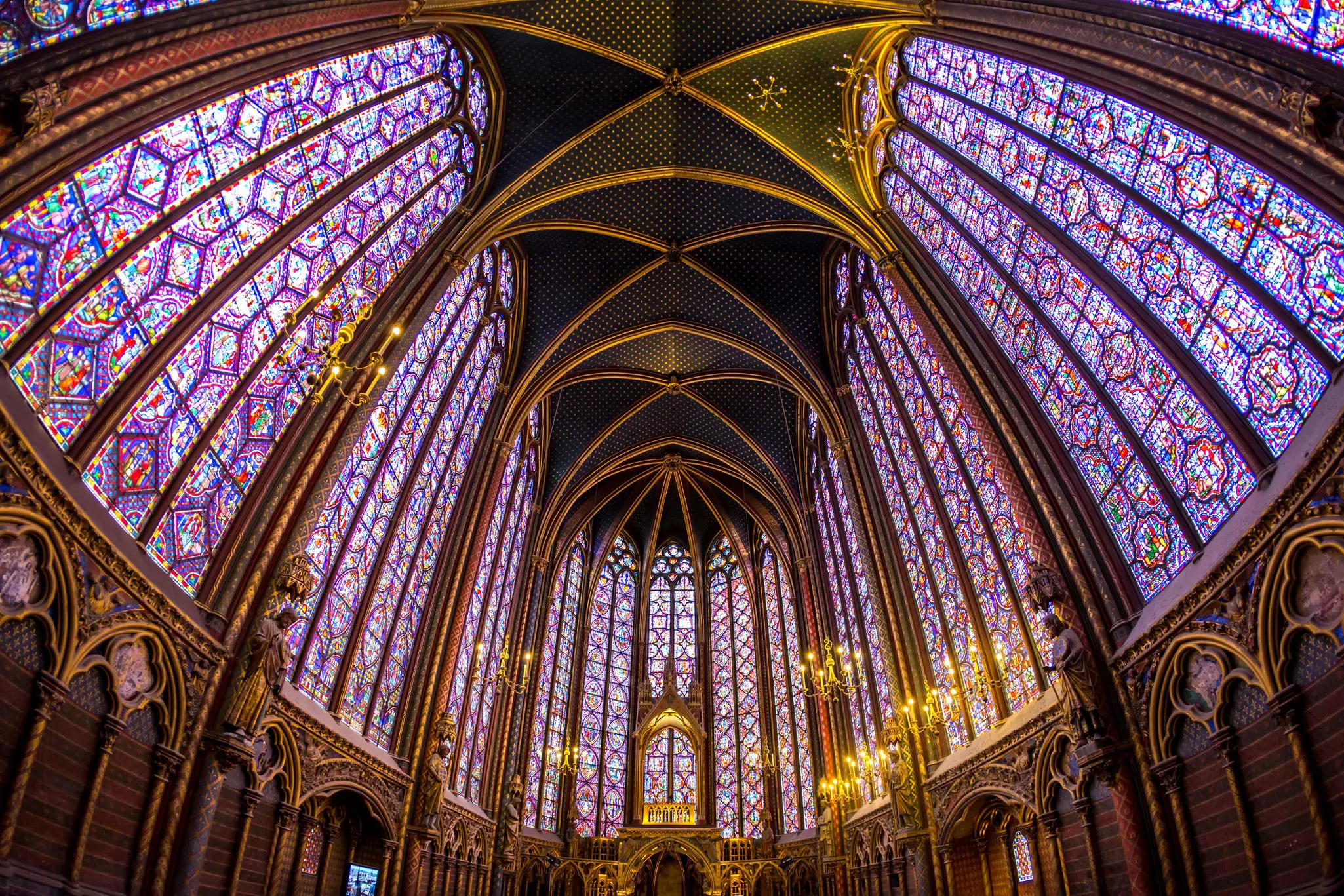 The candle-lit interior of a church with many tall stained-glass windows in shades of red and blue
