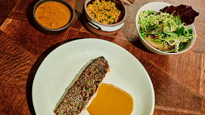 A white plate with a serving of encrusted black bass with an orange sauce sits on a wooden table. Three bowls with beans, yellow rice and green salad are above the white plate.