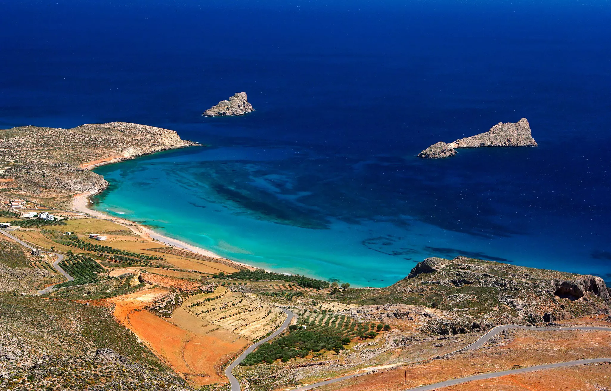 A winding road down a hill leads to a beach, and bright blue waters beyond. Planted groves of olives are visible onshore, with rocky islets in the water.