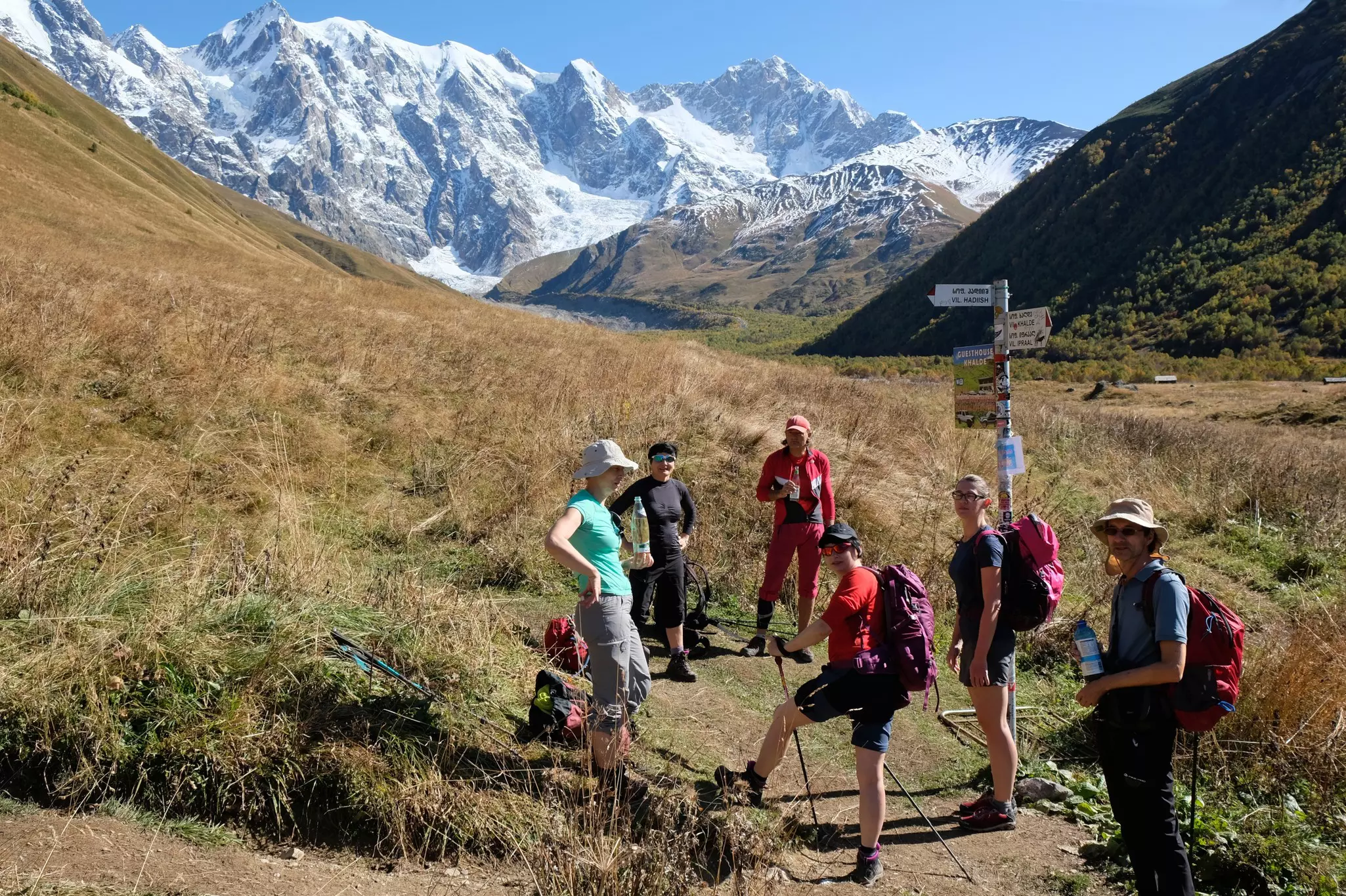 A group of hikers stands in a meadow by a post marking trails. Hills rise around them, with snowy mountains in the distance.