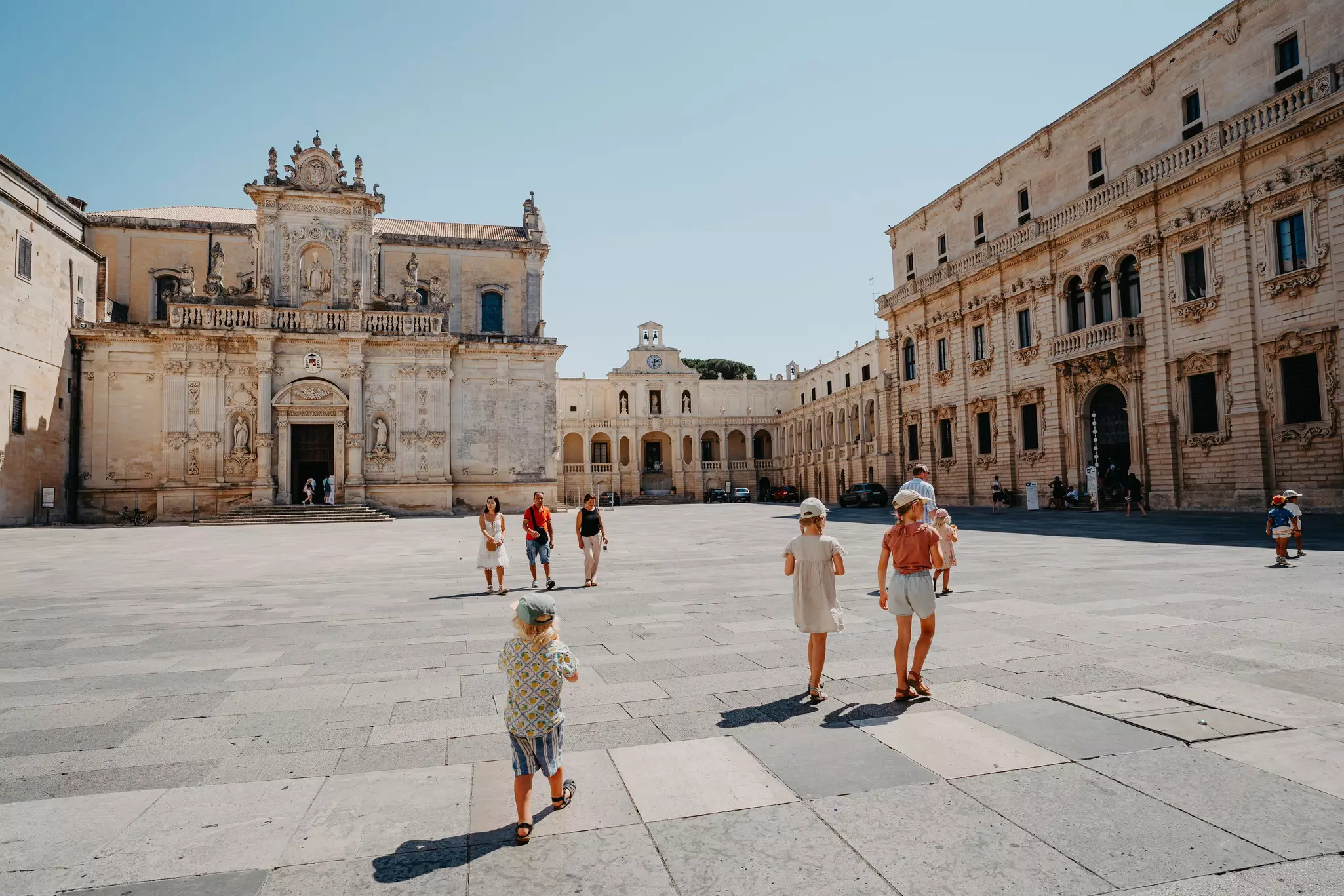 A family walks through a large square of a city on a hot day. A baroque church and other buildings frame the square.