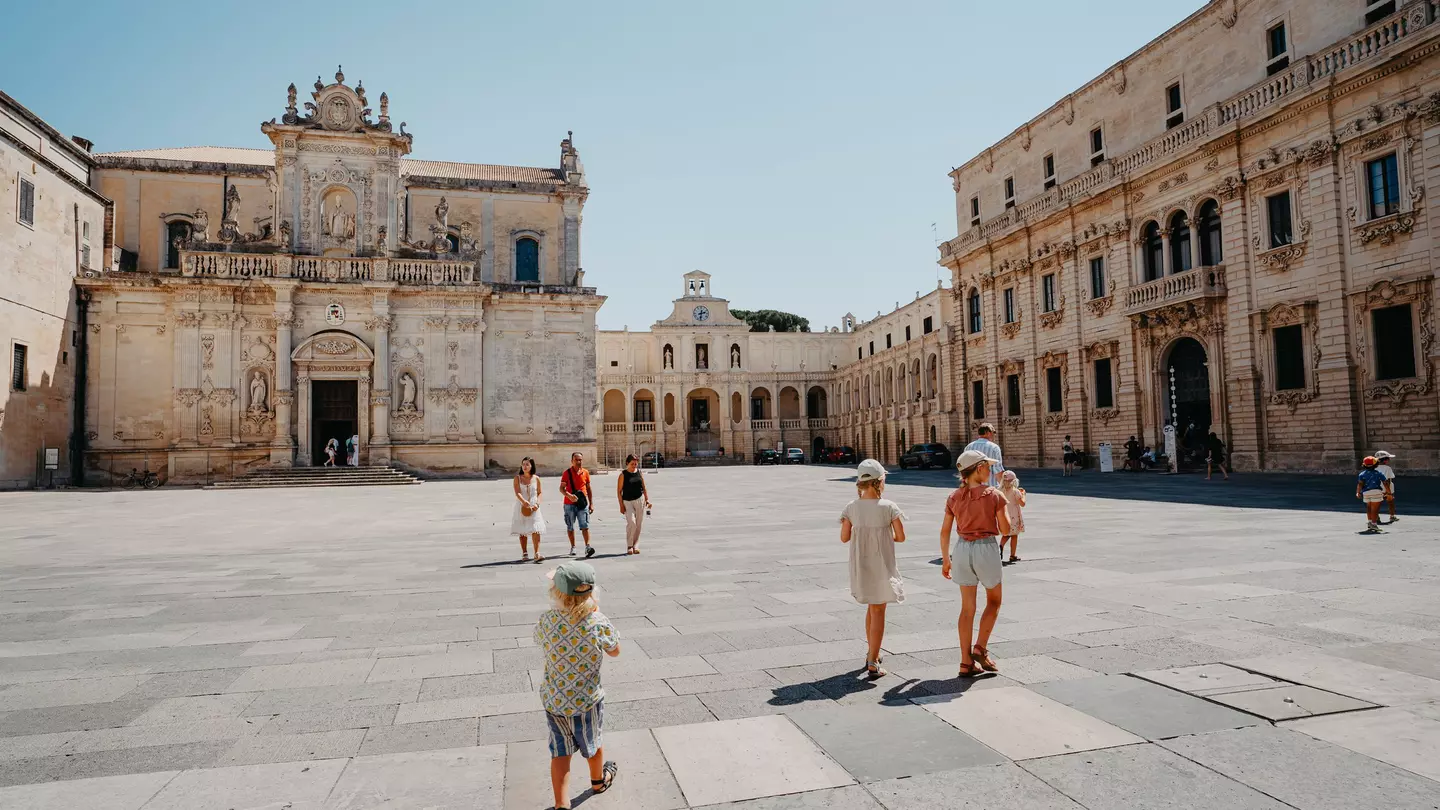 A family walks through a large square of a city on a hot day. A baroque church and other buildings frame the square.