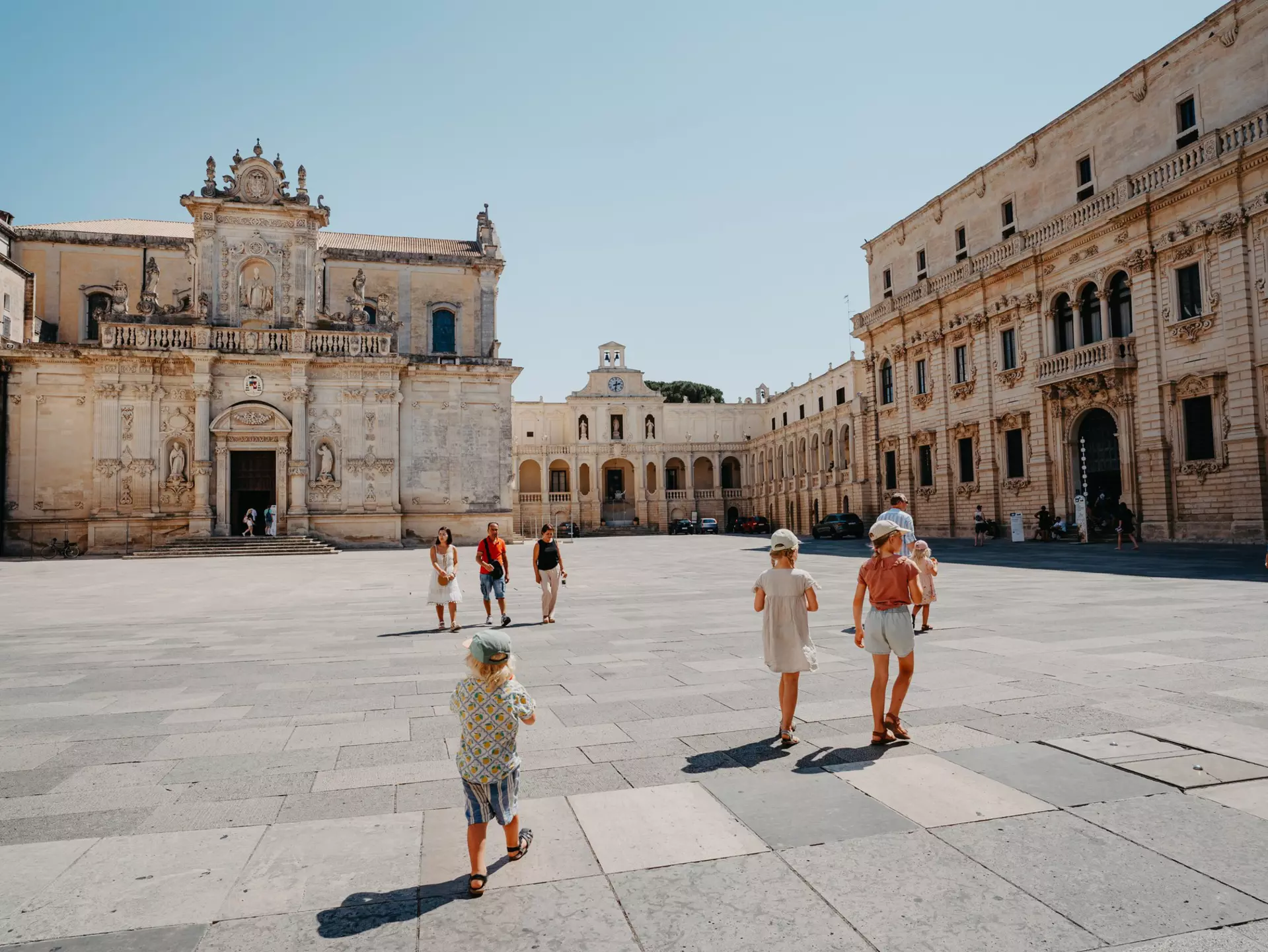 A family walks through a large square of a city on a hot day. A baroque church and other buildings frame the square.
