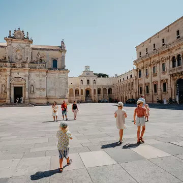 A family walks through a large square of a city on a hot day. A baroque church and other buildings frame the square.