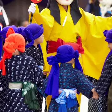 Young people in patterned, blue robelike clothing and colorful headwraps and sashes surround a person in a yellow robe with a red sash. The person is wearing a large mask and carrying what appears to be a yellow fan with red trim and  a large red dot