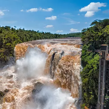 A wide shot of a waterfall swollen with rushing water. Green jungle trees are seen on either side of the waterfall.