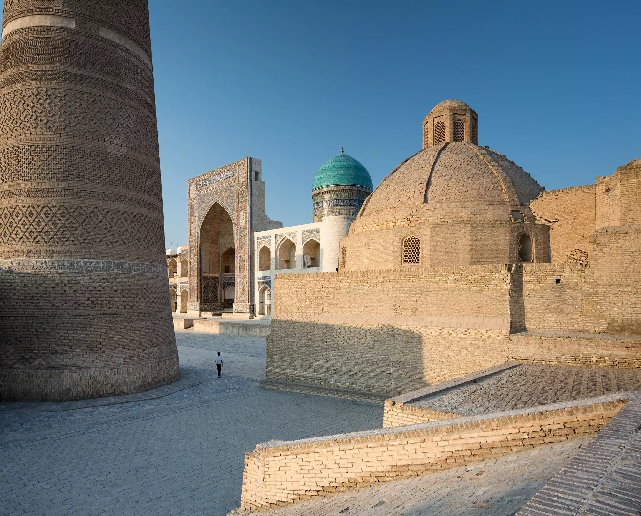 A solo figure passes a brick minaret leading to an ancient archway in Bukhara, Uzbekistan.