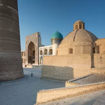 A solo figure passes a brick minaret leading to an ancient archway in Bukhara, Uzbekistan.