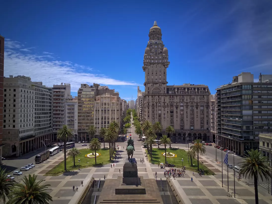 Ornate domed tower on a wide stone building beyond a palm-lined square with an equestrian statue and parked buses