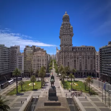 Ornate domed tower on a wide stone building beyond a palm-lined square with an equestrian statue and parked buses