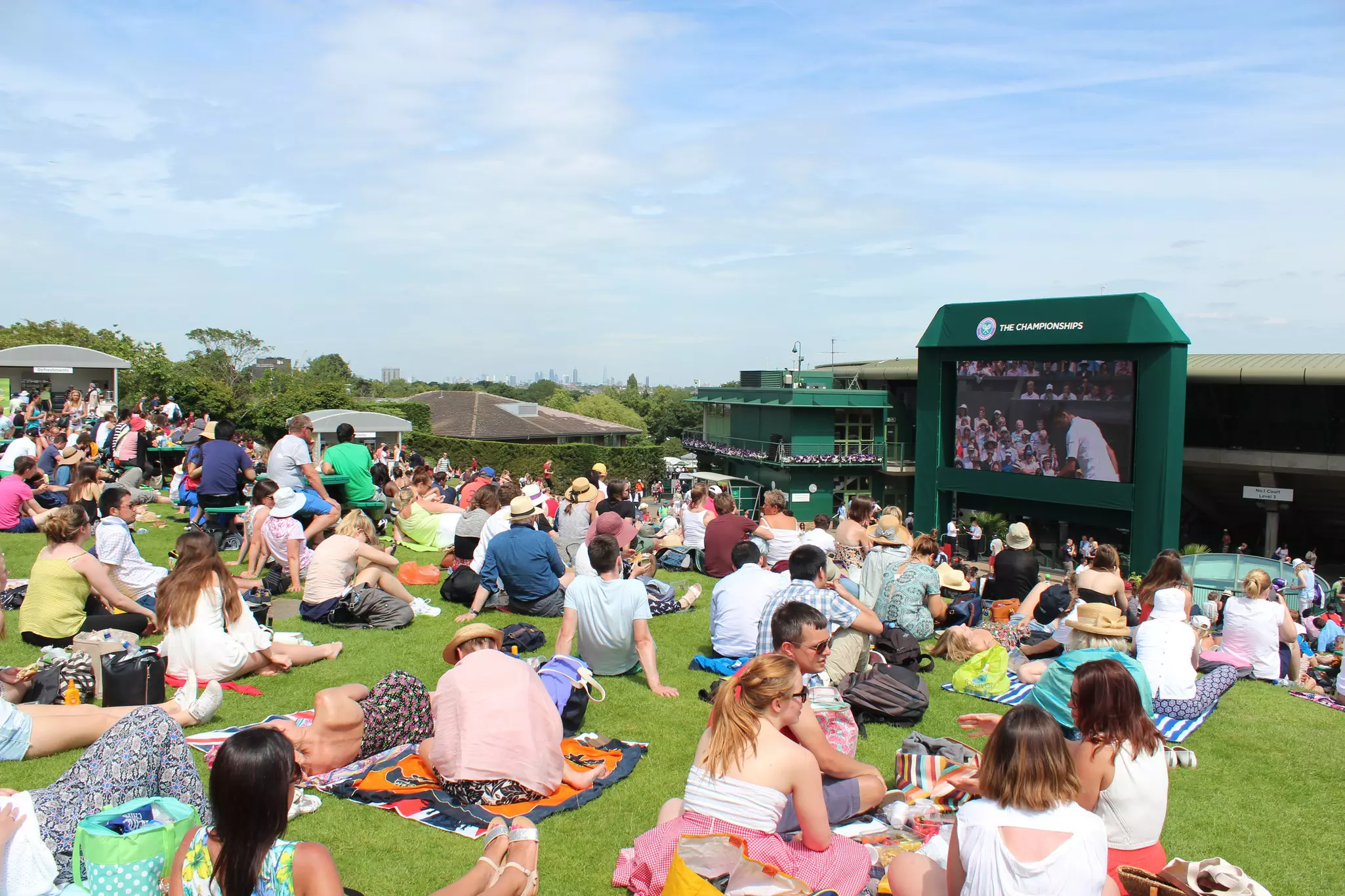 A crowd sits on the grass to watch Wimbledon tennis tournament on a large screen.