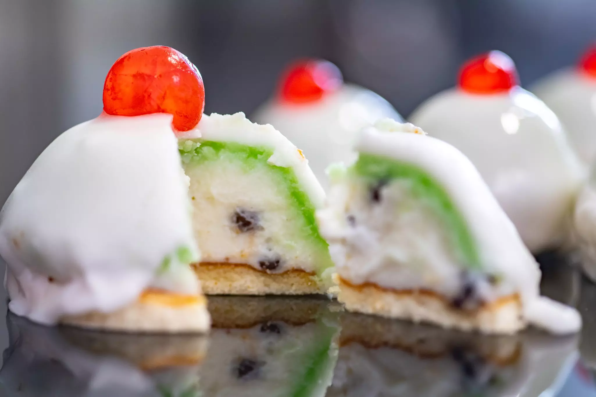 A cross section of a dome-shaped dessert with a creamy center on a thin cookie, which is surrounded by layers of green and white icing, with a red cherry on top.