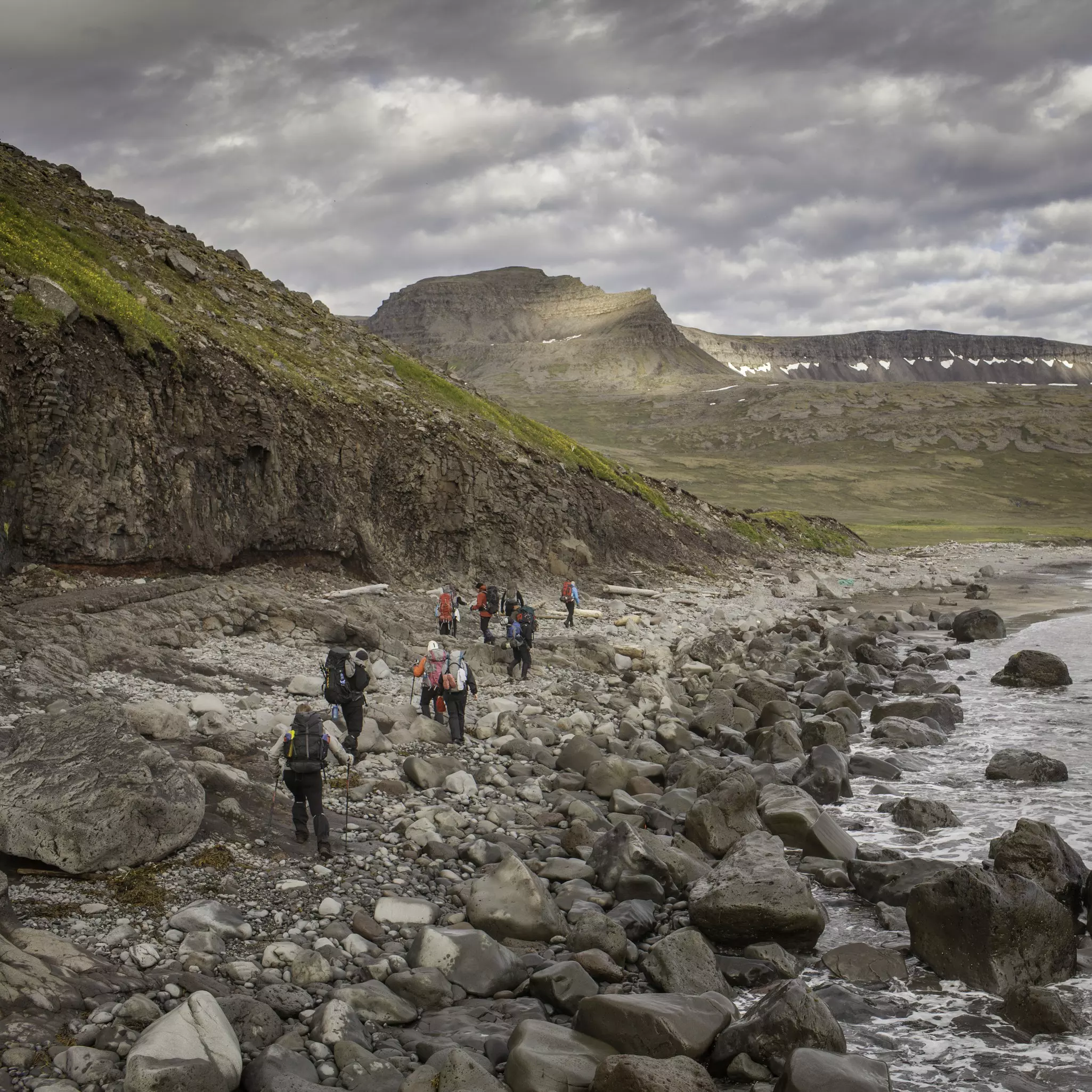A group of hikers walking along a rocky shore, with mountains in the distance