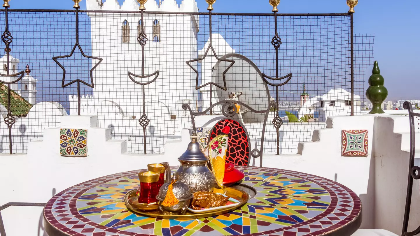 A silver teapot and bowl sit on a tray set on a mosaic round table; a white structure is visible in the background.