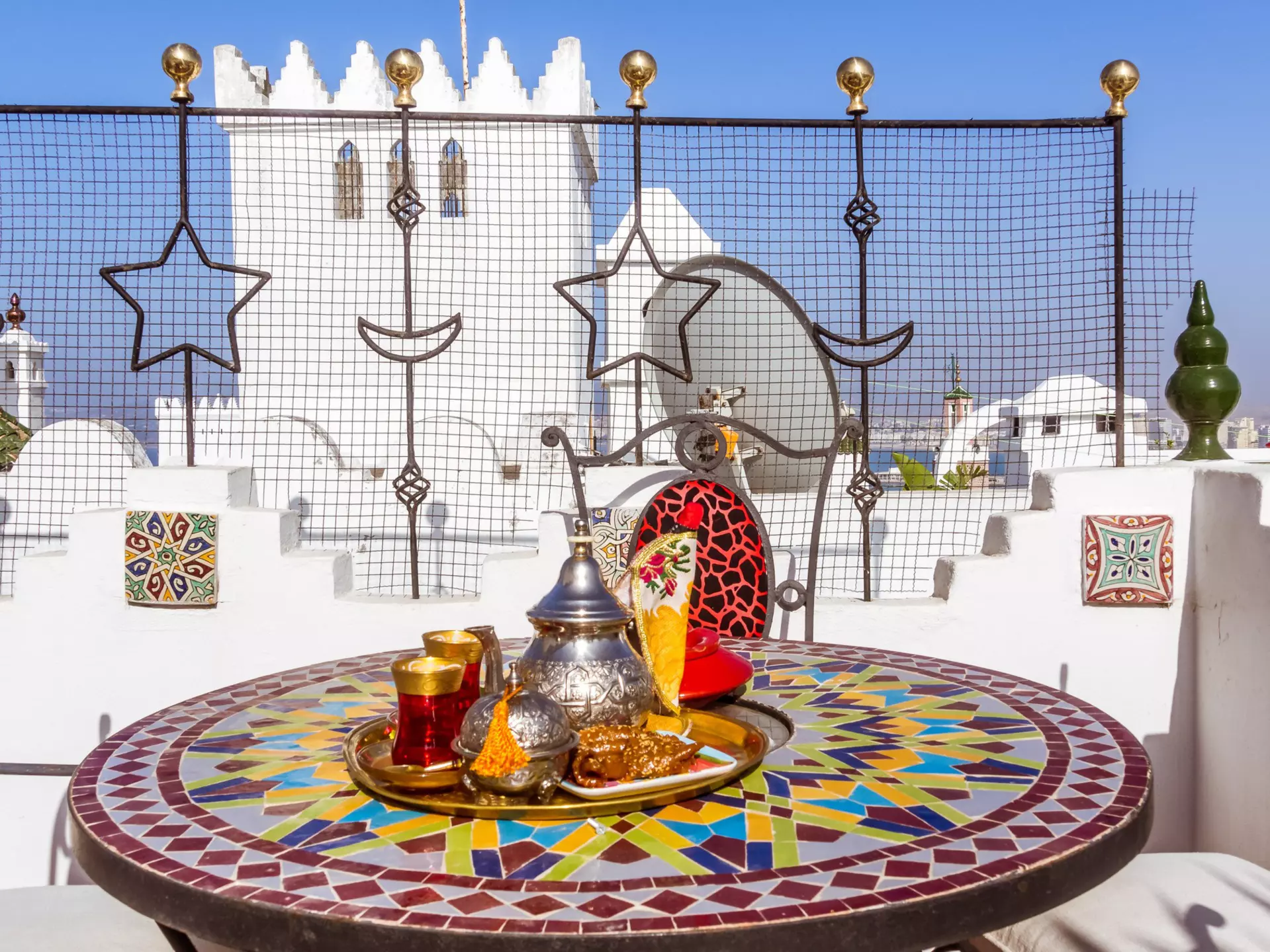 A silver teapot and bowl sit on a tray set on a mosaic round table; a white structure is visible in the background.