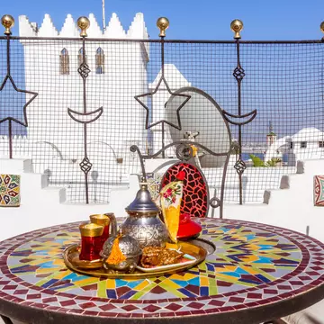 A silver teapot and bowl sit on a tray set on a mosaic round table; a white structure is visible in the background.