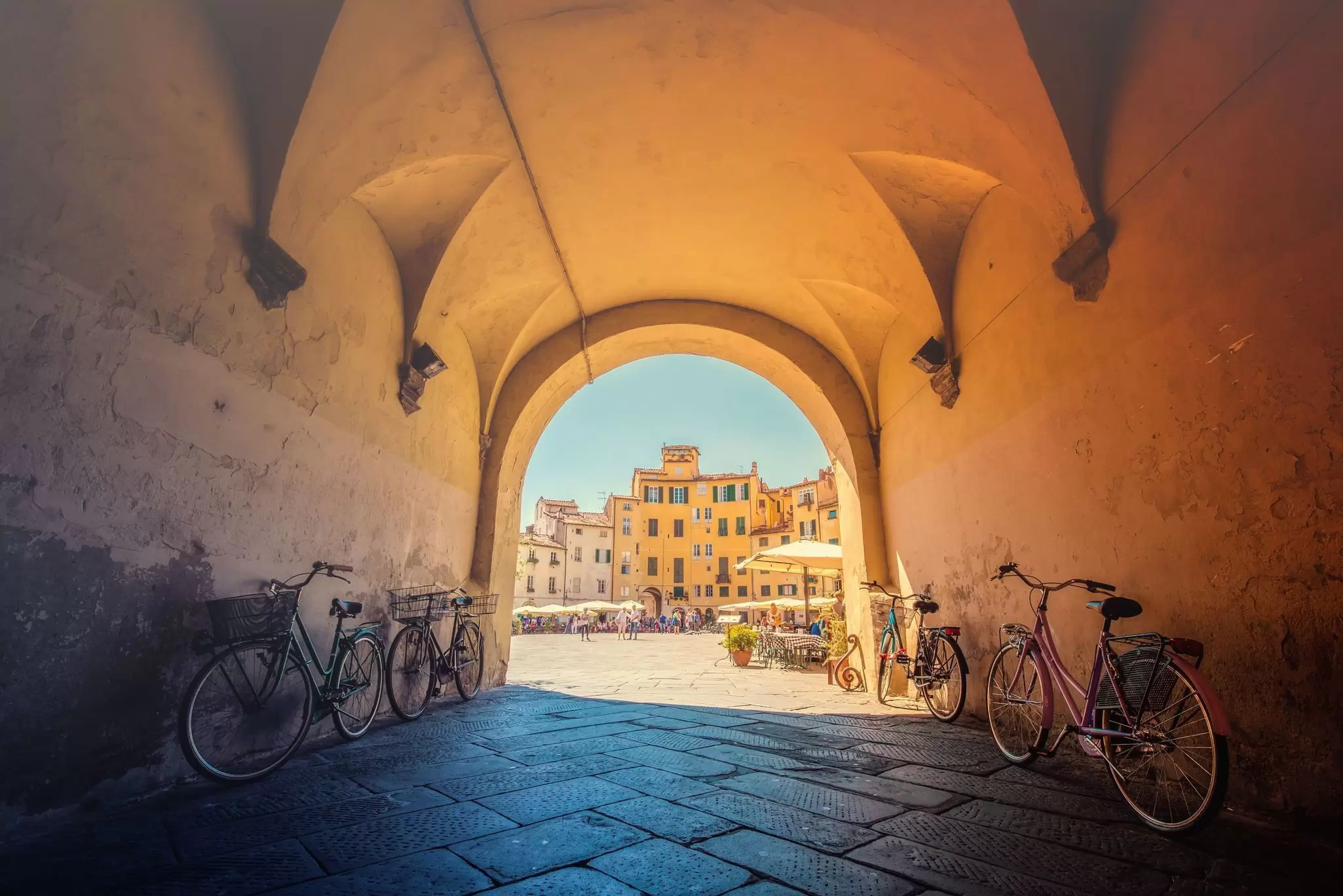 A view of a sun-soaked piazza through a stone tunnel ending in an archway