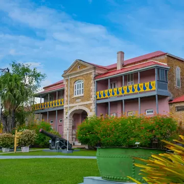 Two-storey stone and pink building with blue and yellow balcony trim and red roof, an old cannon and gardens in front.