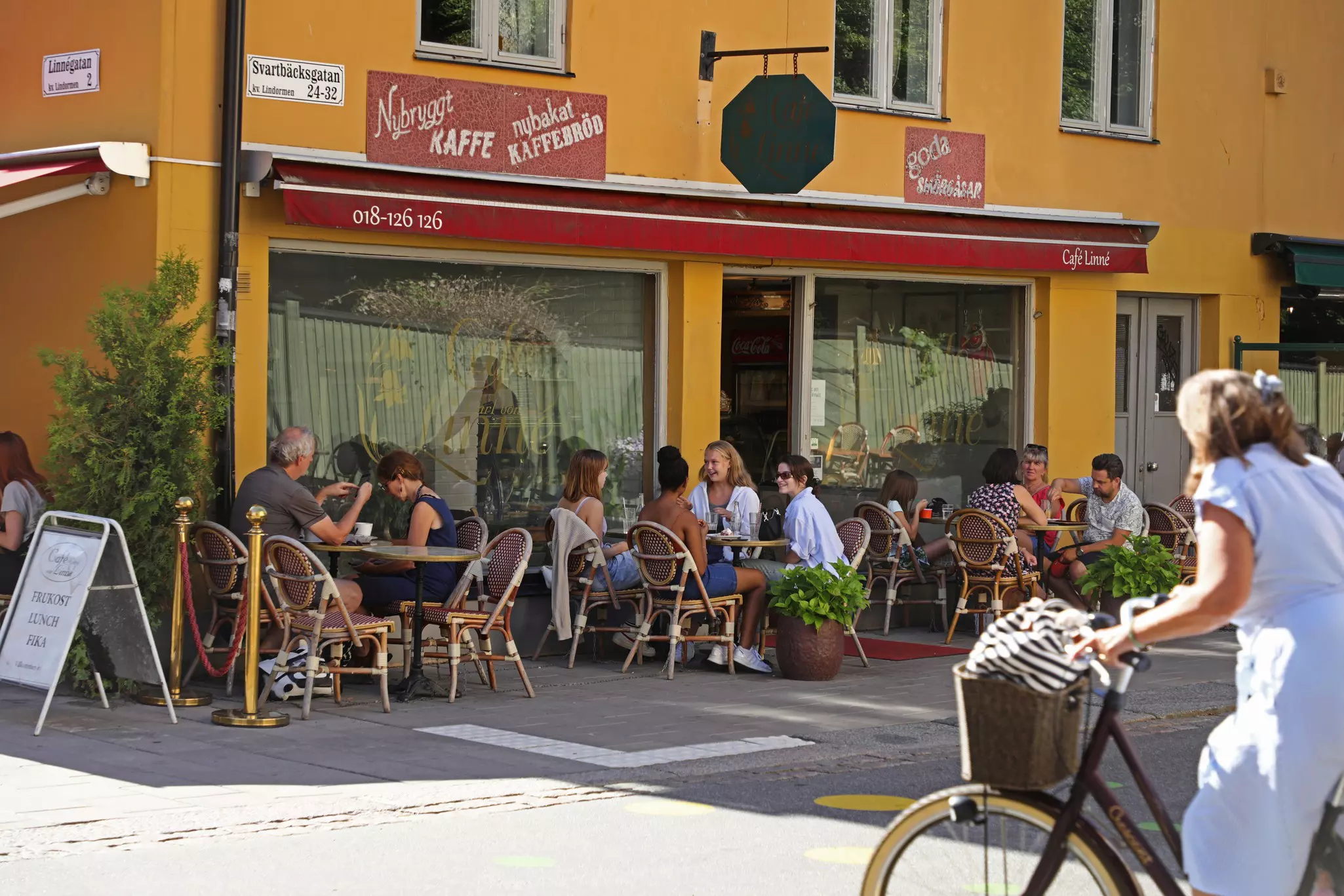 People sit at outdoor tables at a cafe in a city street. A woman walking a bike is seen in the foreground.