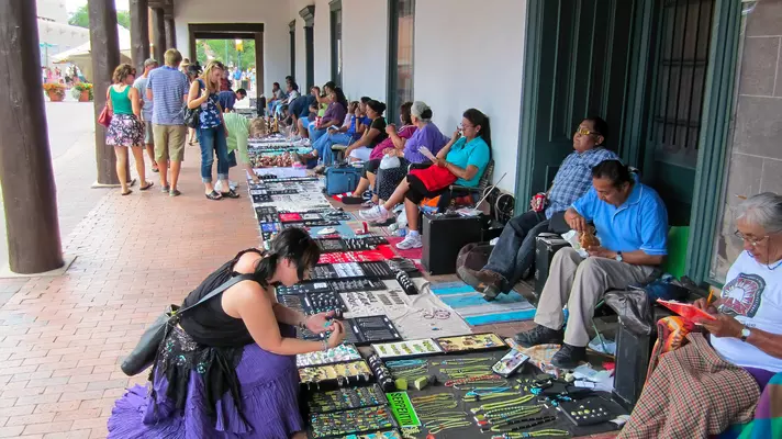 Shoppers look at goods spread on blankets on a walkway under an arcade. Vendors sit in chairs on the other side.