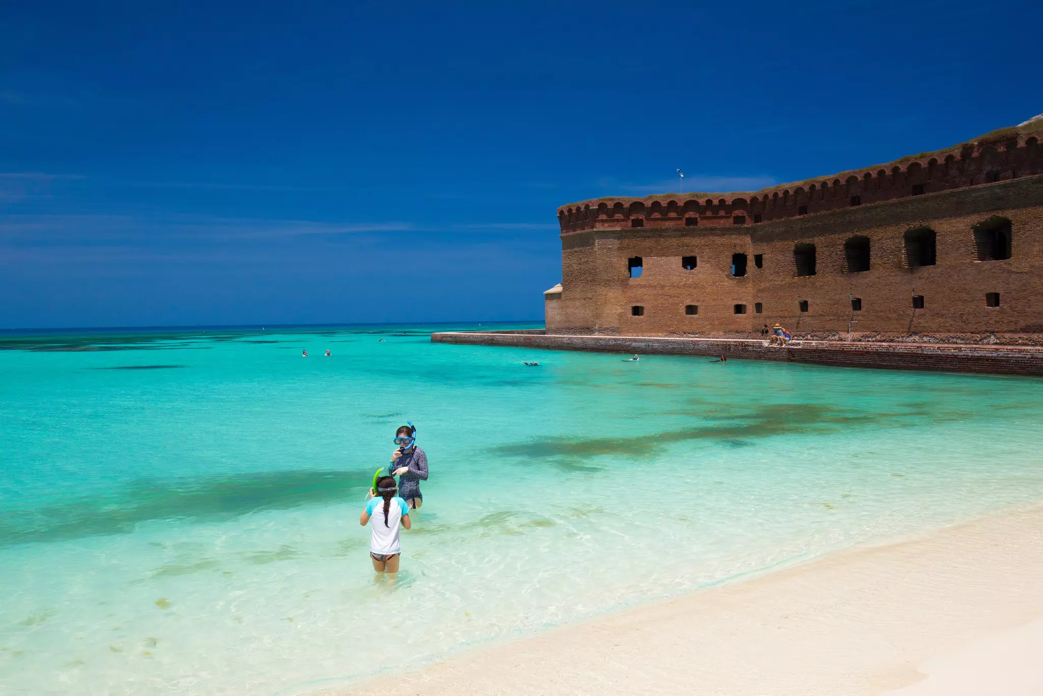 Two people wearing snorkel gear wade into azure waters of a beach. An old fortress is seen behind them.