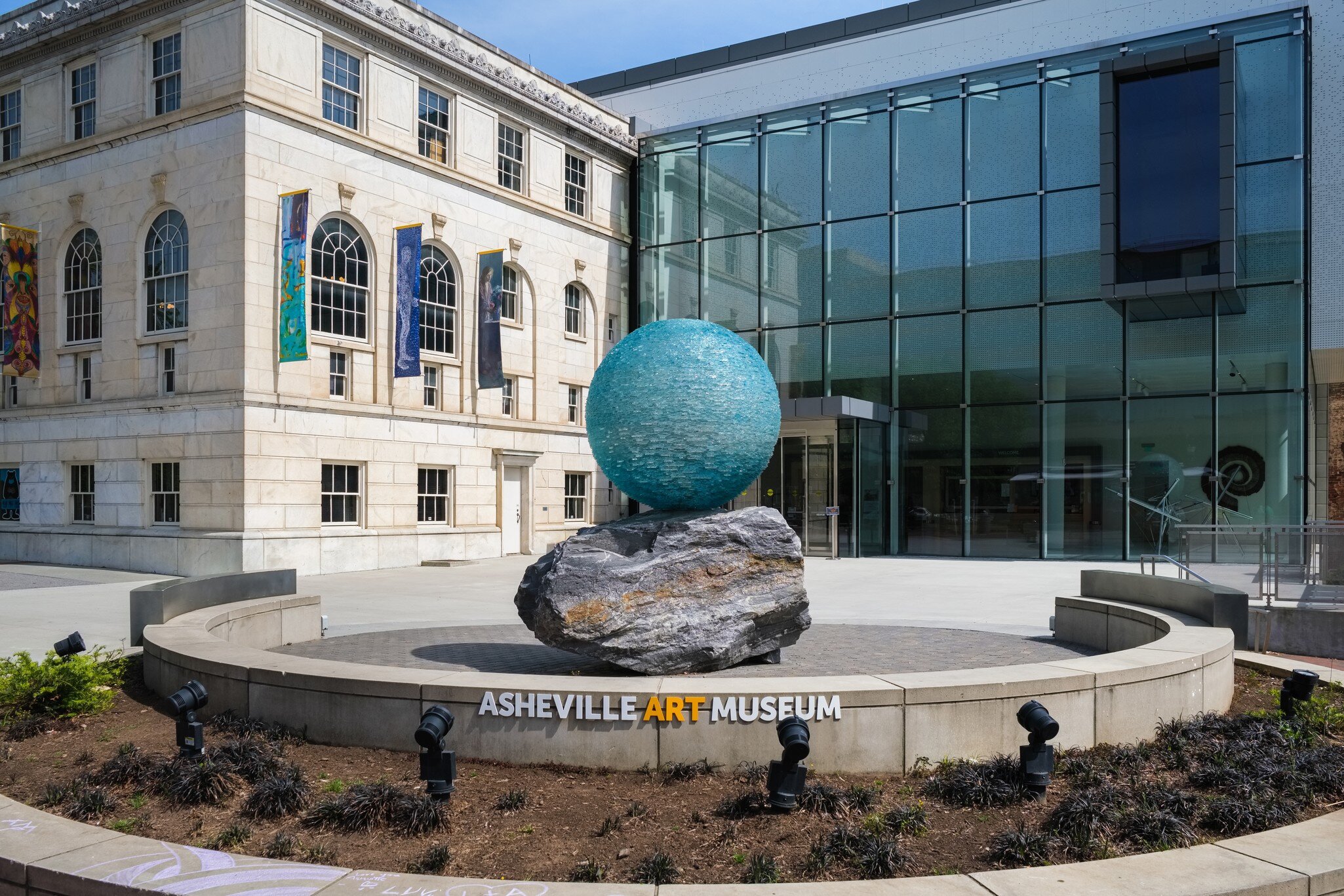 Teal glass sphere on a boulder above a curved wall lettered ASHEVILLE ART MUSEUM, next to a glass atrium wing.