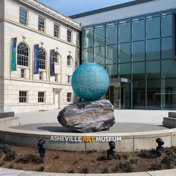 Teal glass sphere on a boulder above a curved wall lettered ASHEVILLE ART MUSEUM, next to a glass atrium wing.