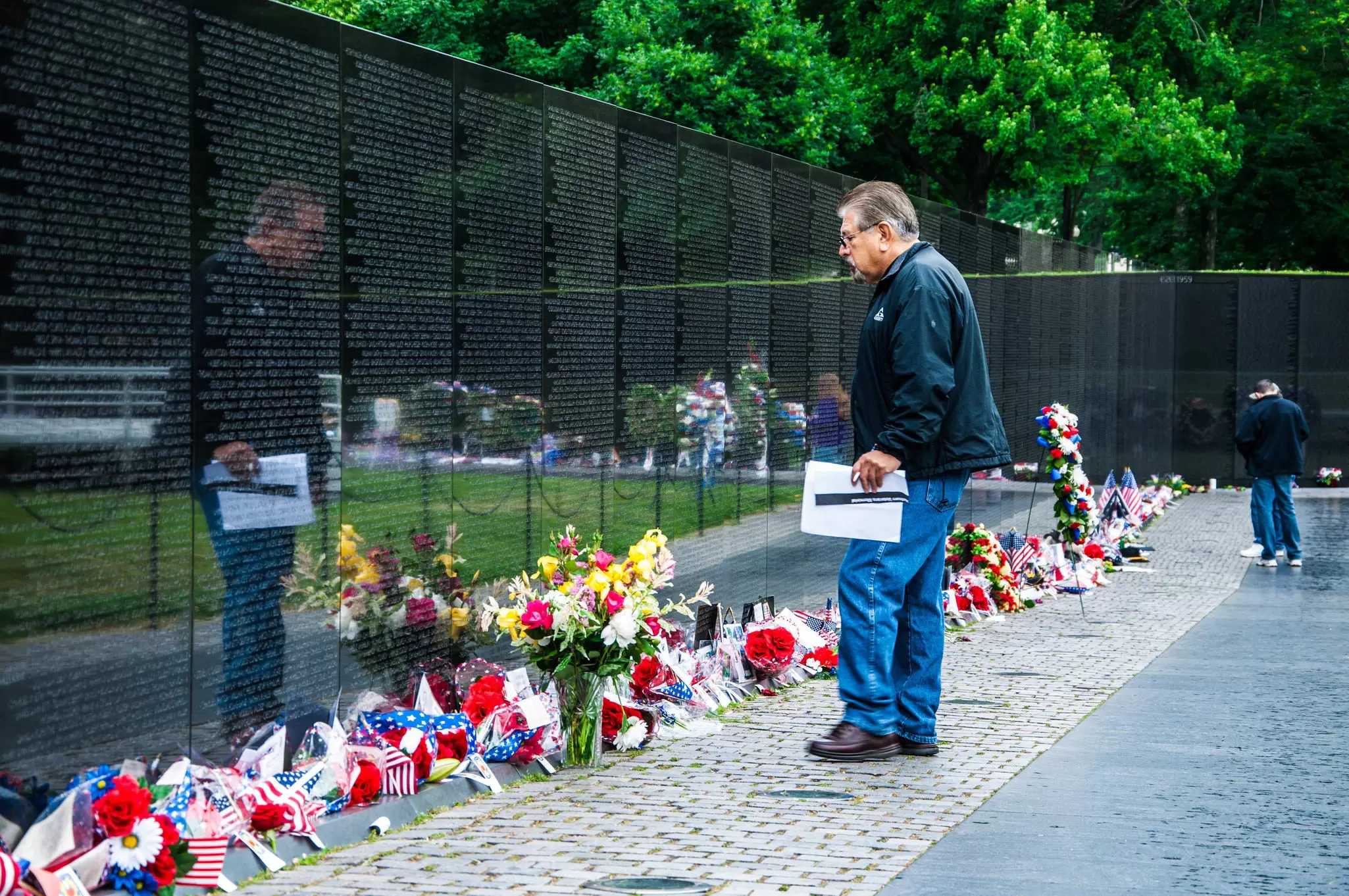 A man stands in front of the memorial. Flowers are all along the front of the wall.