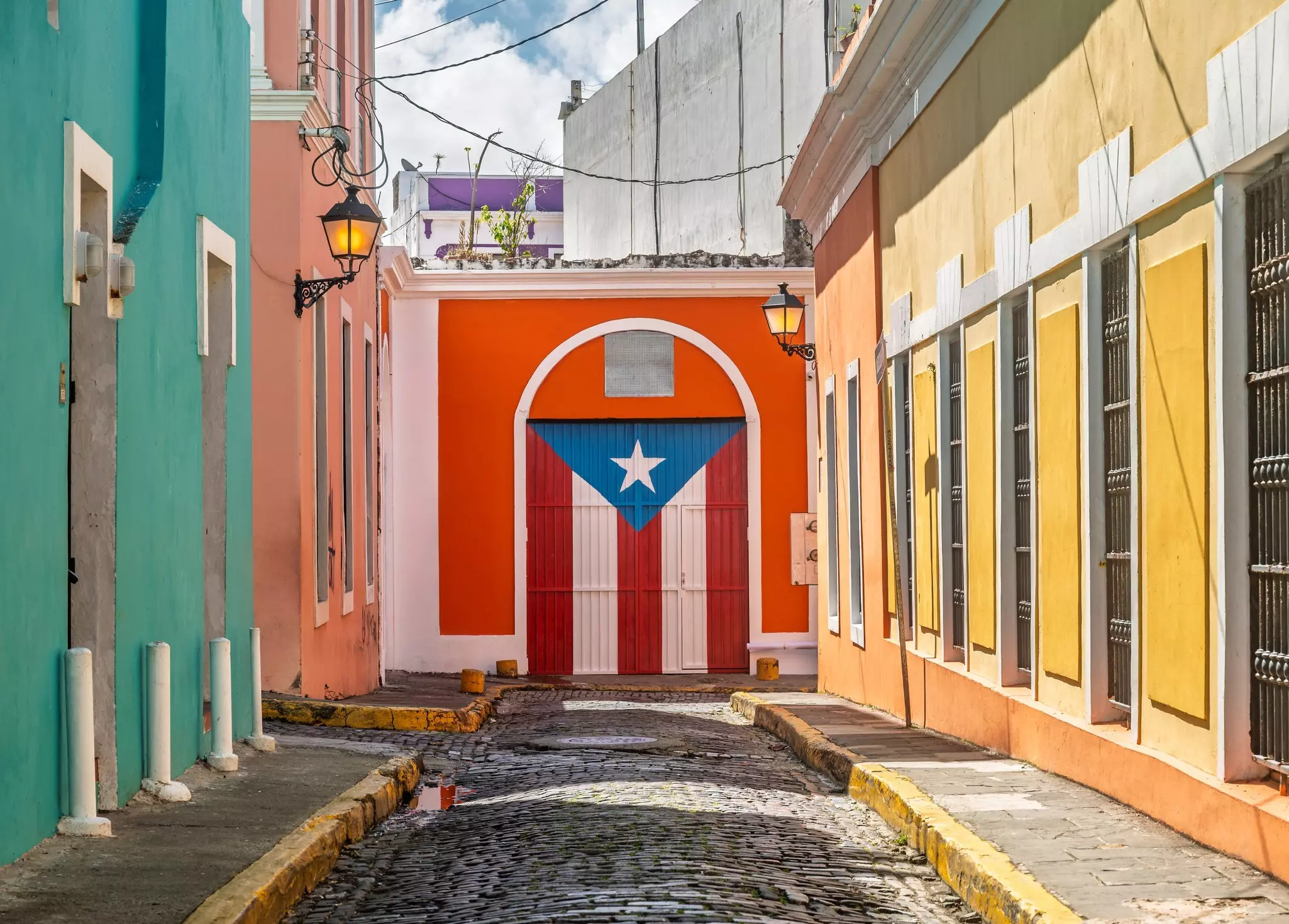 Green, coral and yellow buildings on a narrow cobblestone street that terminates at a doorway painted with the flag of Puerto Rico.