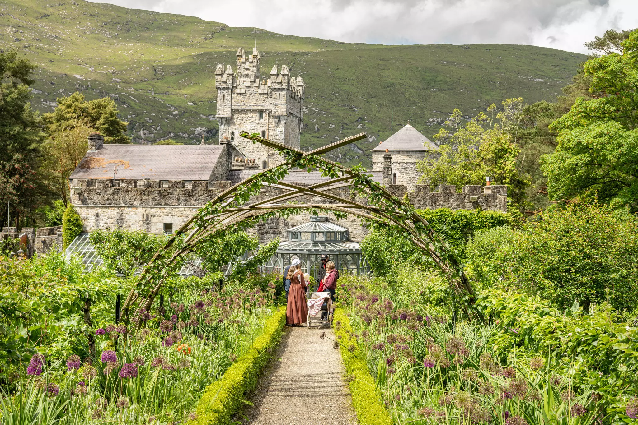 A family stands in a castle garden full of flowering plants with mountains in the distance.