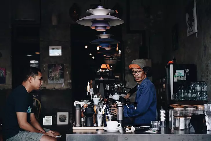 A barista preparing a drink behind a counter in a darkened coffeehouse while a patron waits on the other side