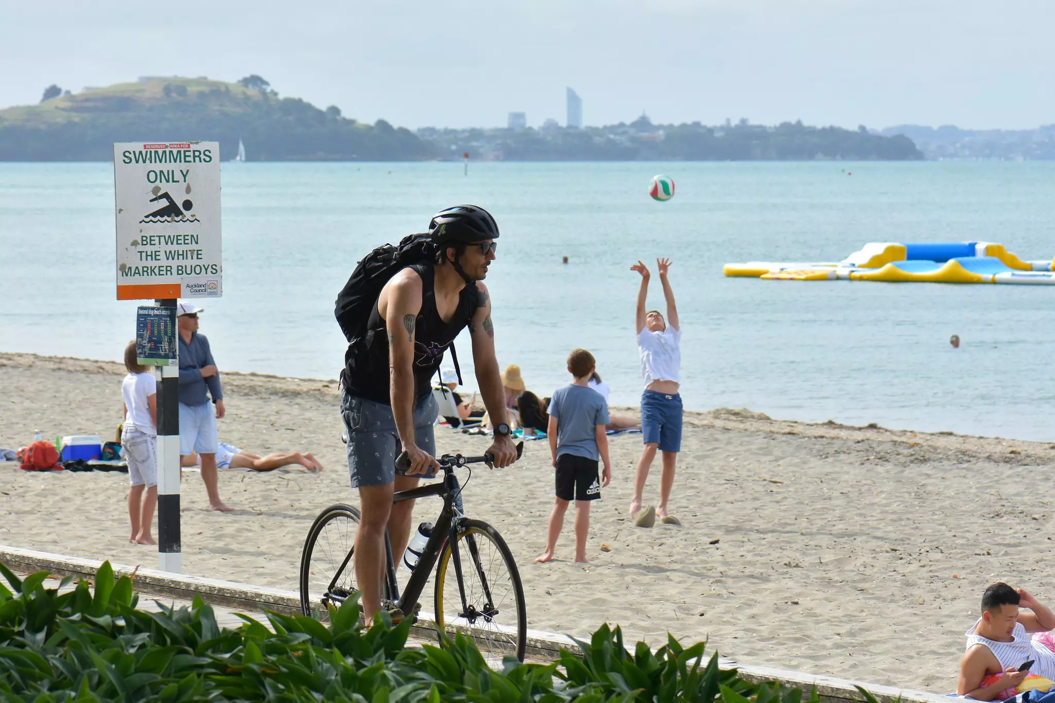 A cyclist rides past a sandy beach where people are playing with a ball.