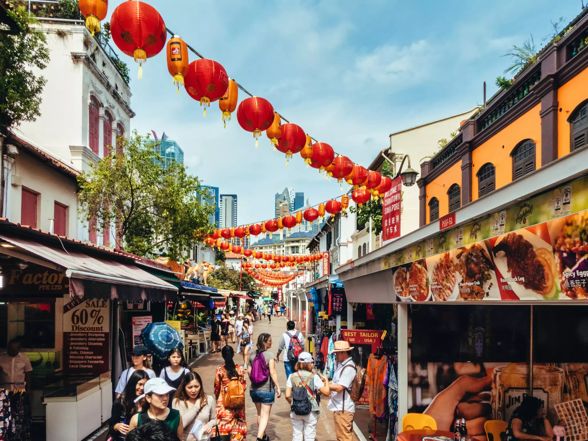 Red lanterns hang over a narrow street filled with pedestrians shopping at market stalls in a city.
